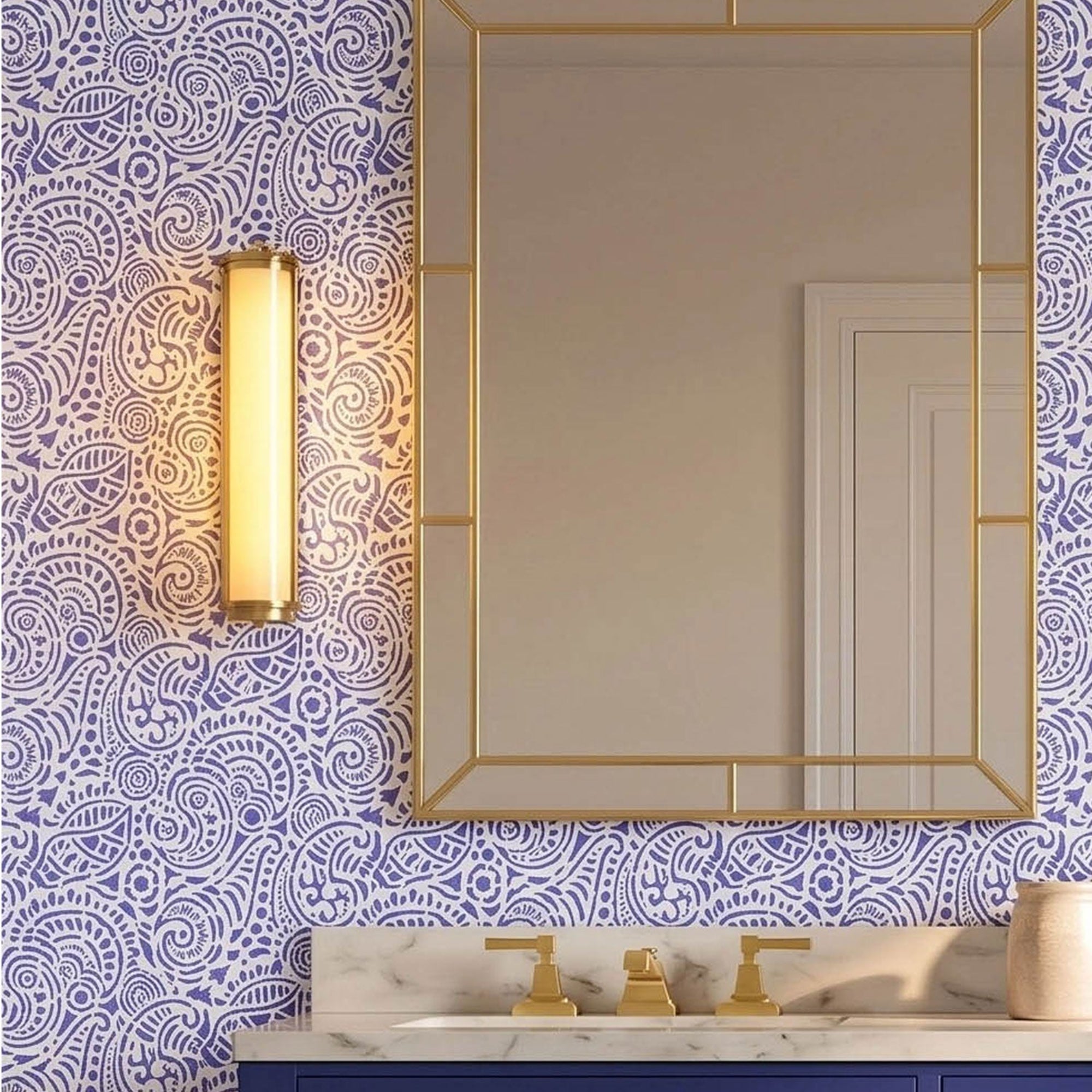 Abstract seashells wall stencil applied in a bathroom vanity with a deep blue cabinet, white marble countertop, gold-framed mirror and wall sconce, and light highlighting the dark blue and white painted stencil on the wall.