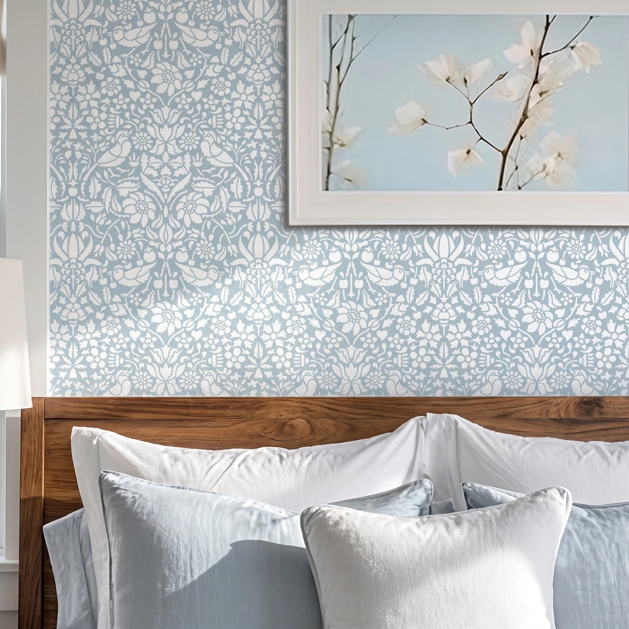 Birds and berries wall stencil applied on a wall above a wooden headboard with white pillows and a framed print of white flowers on a light blue sky and natural light highlighting the blue and white painted stencil on the wall.