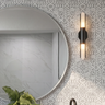 A modern wall-mounted sconce with a black metal frame and two vertical frosted glass lights emitting warm light, mounted on the wall painted with botanical tile wall stencil in grey next to a large round mirror.