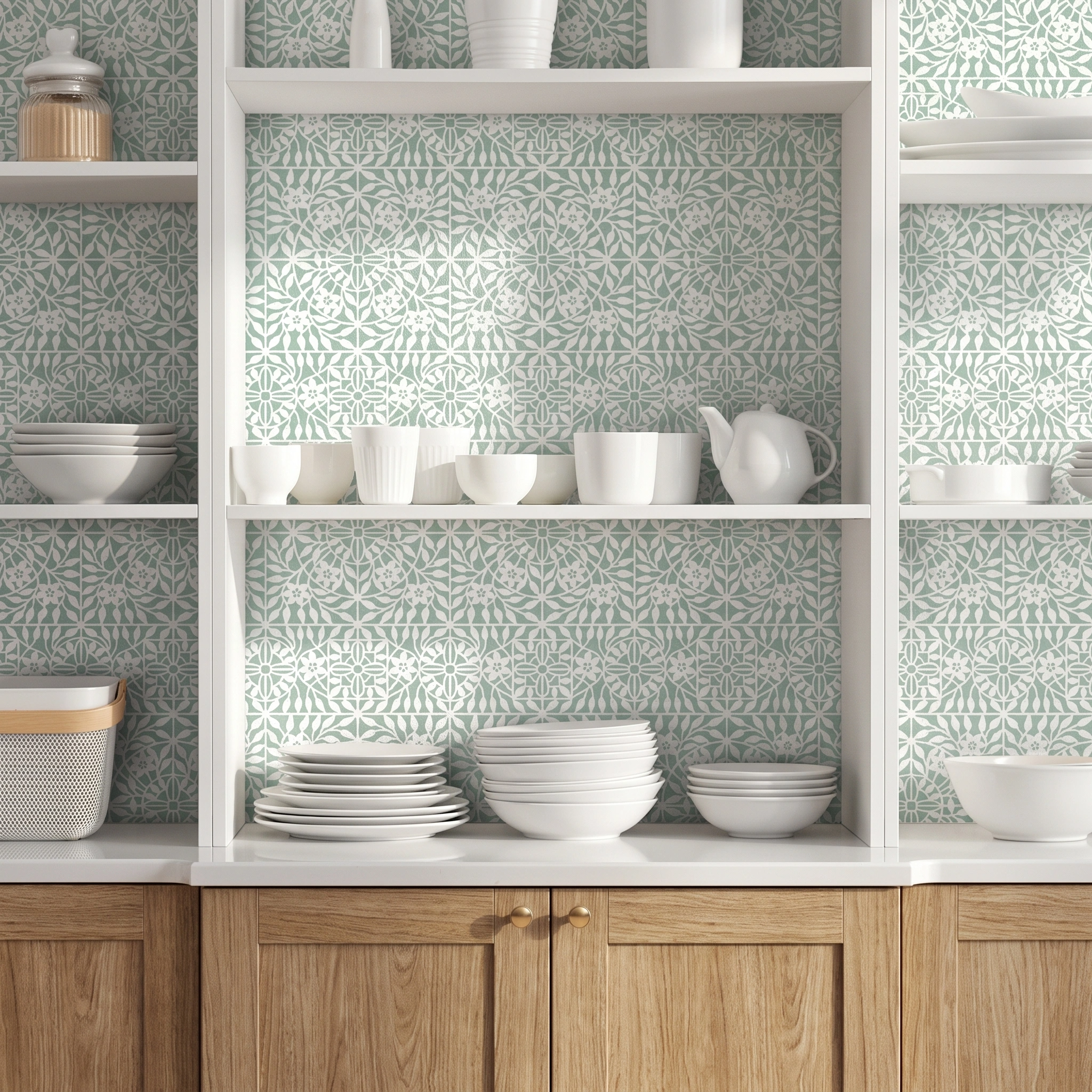 A styled kitchen interior with white open shelving and wooden cabinets, decorated with a repeating light green and white botanical tile wall stencil. White ceramic dishes, bowls, cups, and a teapot are neatly arranged on the shelves.