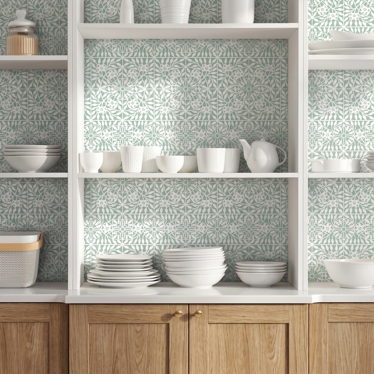 A styled kitchen interior with white open shelving and wooden cabinets, decorated with a repeating light green and white botanical tile wall stencil. White ceramic dishes, bowls, cups, and a teapot are neatly arranged on the shelves.