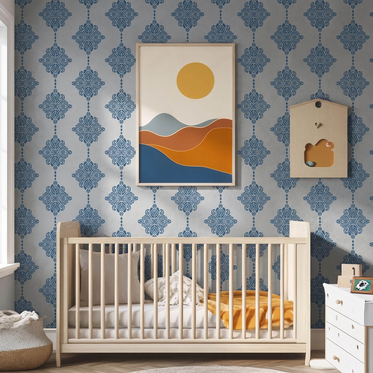 Damask block print stencil applied in a nursery, styled with a minimalist framed art print of rolling hills and a sun, and the top of a light wood crib, with natural light highlighting the dark blue on light blue stencil painted on the wall.