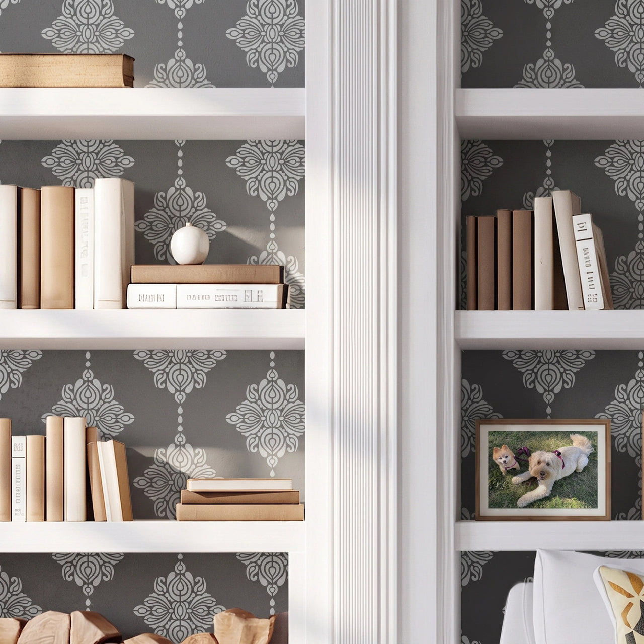 Damask block print wall stencil applied behind a white built-in bookshelves, books, firewood stack, and a white armchair with natural light highlighting the dark gray and light gray paint stencil painted on the wall behind the built-in shelves.