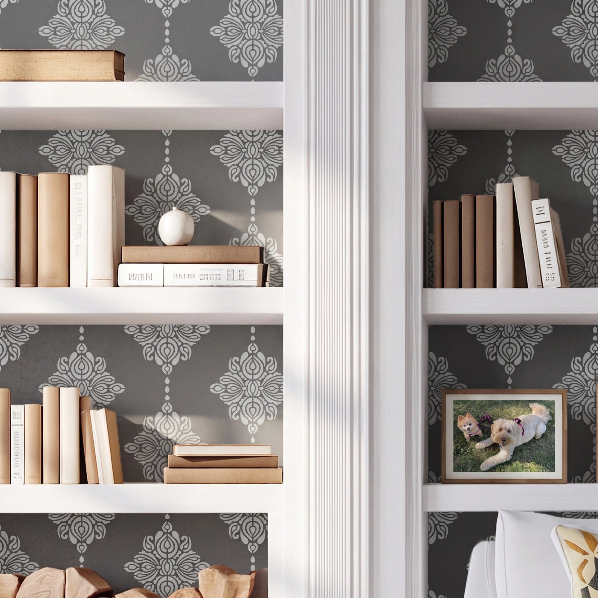 Damask block print wall stencil applied behind a white built-in bookshelves, books, firewood stack, and a white armchair with natural light highlighting the dark gray and light gray paint stencil painted on the wall behind the built-in shelves.
