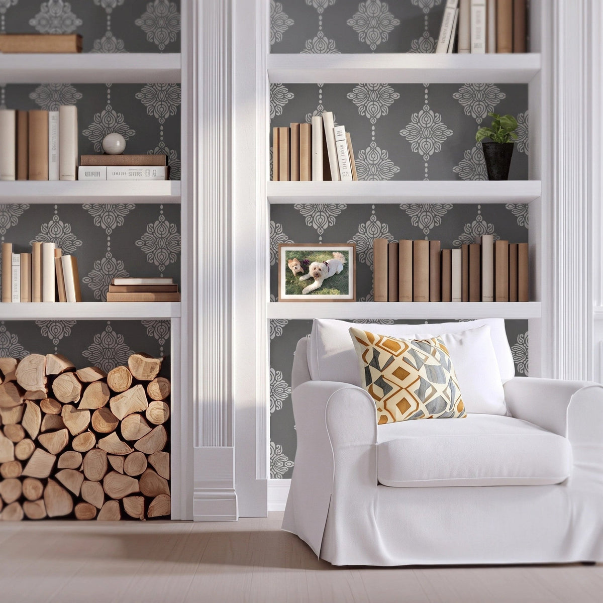Damask block print wall stencil applied behind a white built-in bookshelves, books, firewood stack, and a white armchair with natural light highlighting the dark gray and light gray paint stencil painted on the wall behind the built-in shelves.