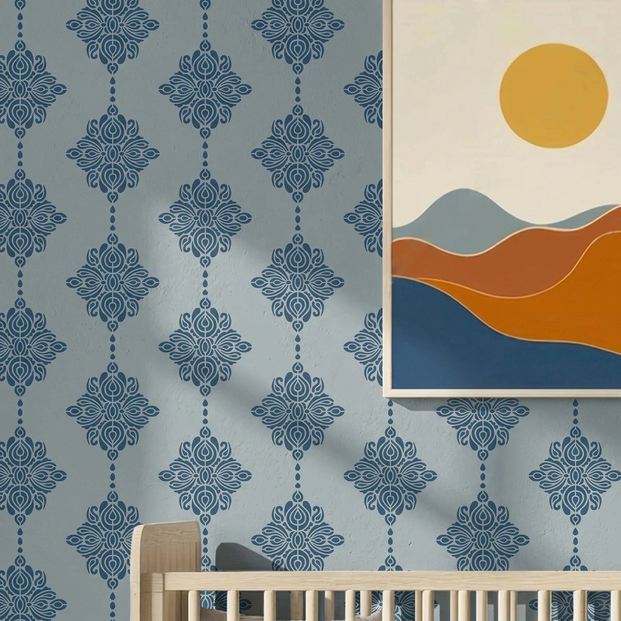 Damask block print stencil applied in a nursery, styled with a minimalist framed art print of rolling hills and a sun, and the top of a light wood crib, with natural light highlighting the dark blue on light blue stencil painted on the wall.