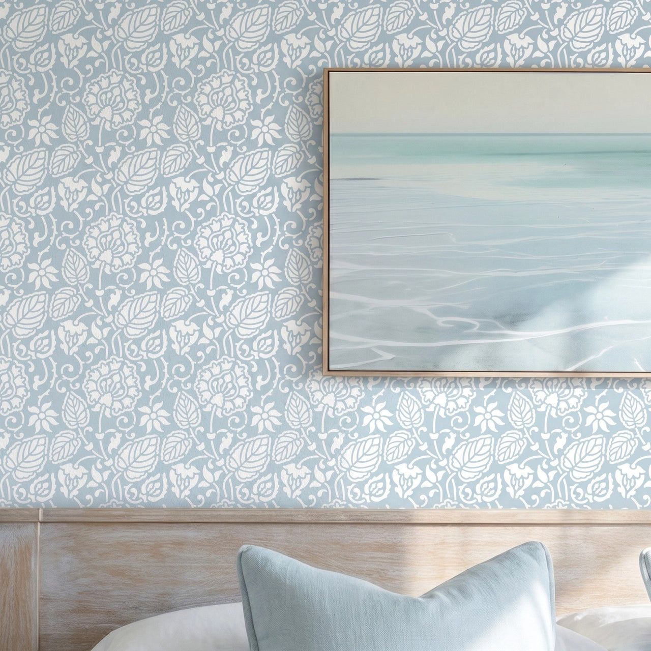 Dandelion wall stencil in bedroom with a neatly made bed in white linens and pale blue pillows, framed abstract seascape print on the wall, and natural light highlighting the blue and white stencil painted on the wall.