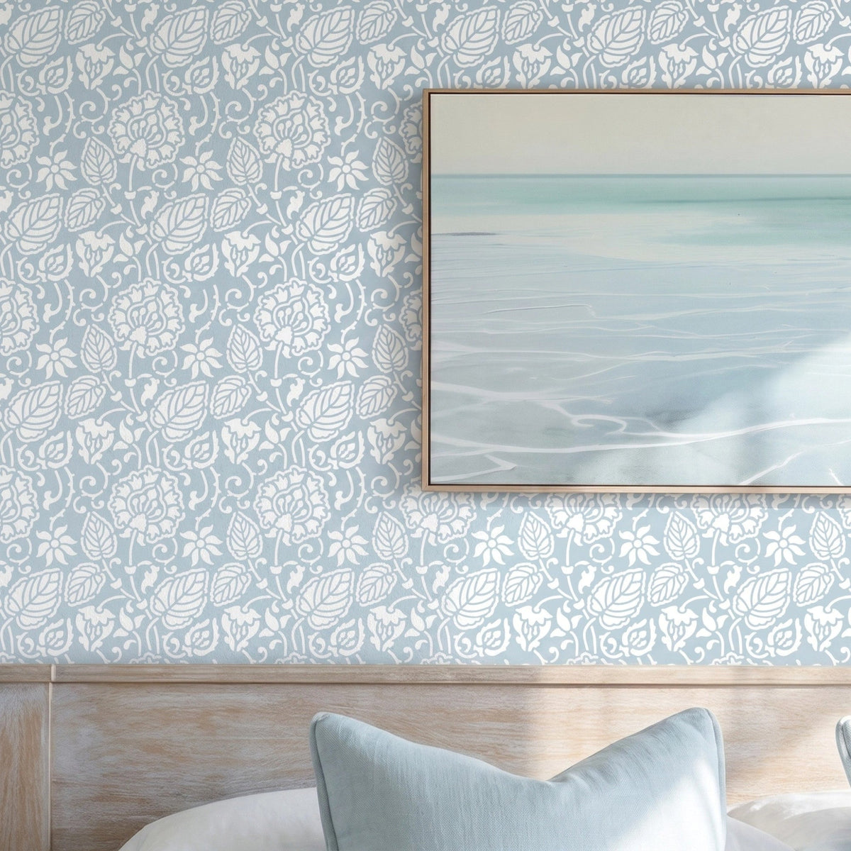 Dandelion wall stencil in bedroom with a neatly made bed in white linens and pale blue pillows, framed abstract seascape print on the wall, and natural light highlighting the blue and white stencil painted on the wall.