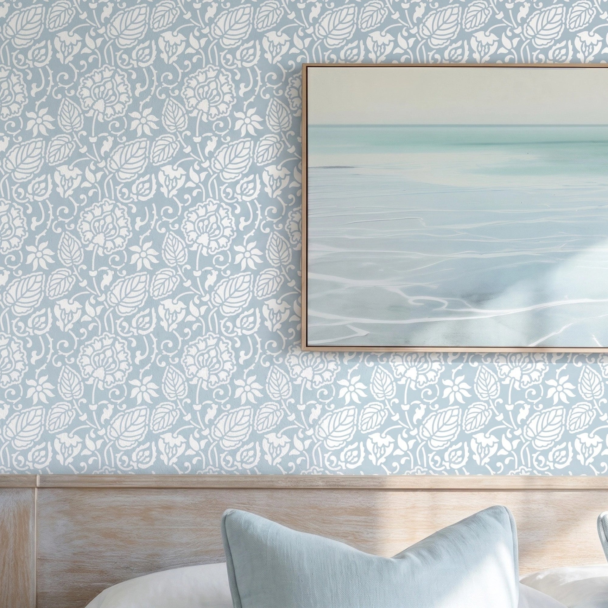 Dandelion wall stencil in bedroom with a neatly made bed in white linens and pale blue pillows, framed abstract seascape print on the wall, and natural light highlighting the blue and white stencil painted on the wall.
