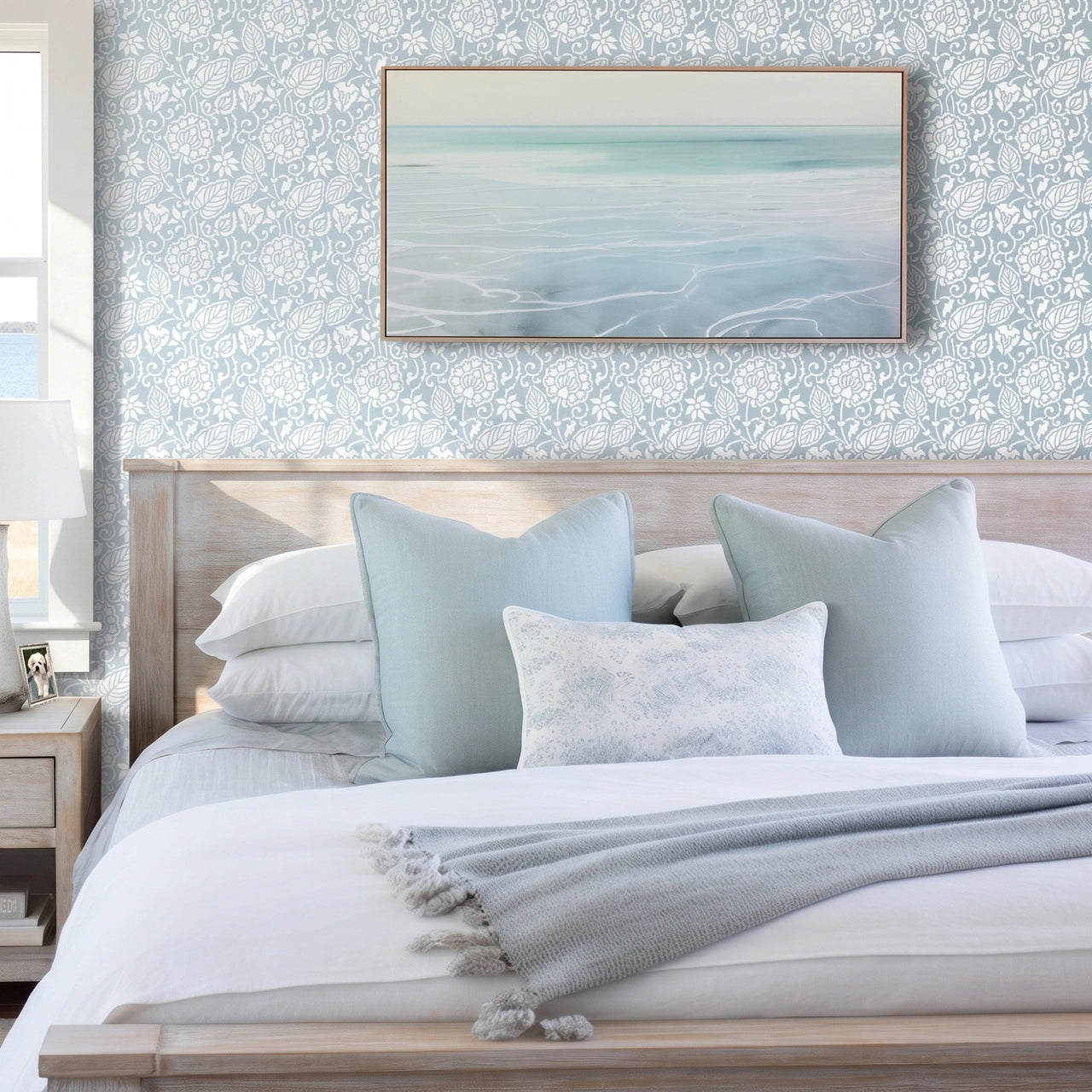 Dandelion wall stencil in bedroom with a neatly made bed in white linens and pale blue pillows, framed abstract seascape print on the wall, and natural light highlighting the blue and white stencil painted on the wall.