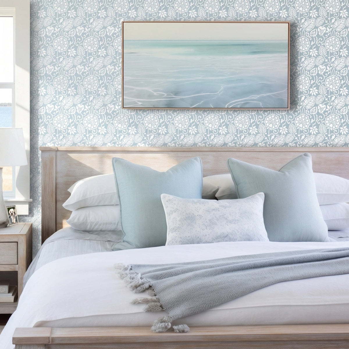 Dandelion wall stencil in bedroom with a neatly made bed in white linens and pale blue pillows, framed abstract seascape print on the wall, and natural light highlighting the blue and white stencil painted on the wall.