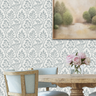 A styled dining room showing a wall painted with floral gallery wall stencil in a muted gray and a off-white background, with part of a framed landscape painting, a wooden dining table, chairs, and a vase of pink flowers.