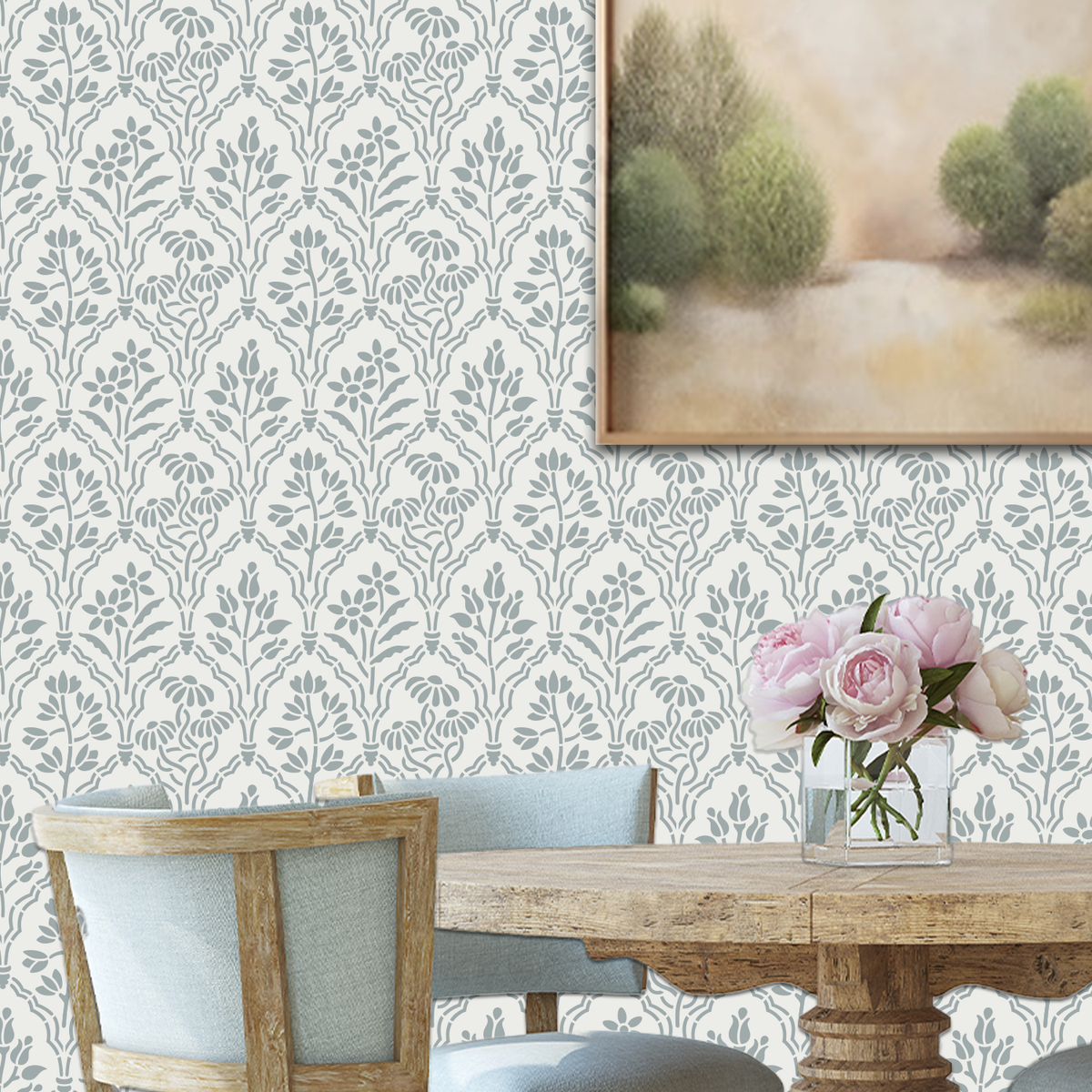 A styled dining room showing a wall painted with floral gallery wall stencil in a muted gray and a off-white background, with part of a framed landscape painting, a wooden dining table, chairs, and a vase of pink flowers.