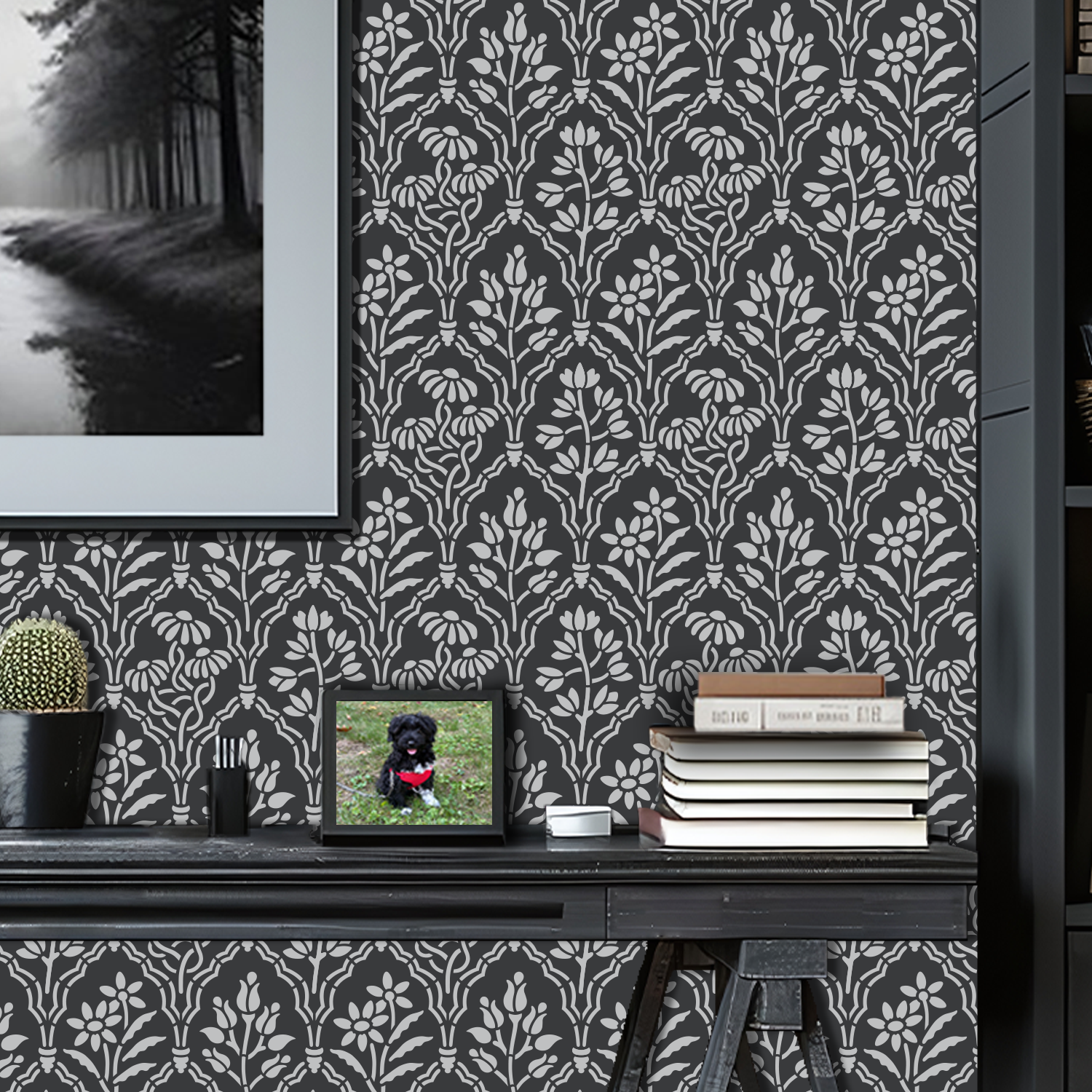 A styled home office scene featuring floral gallery wall stencil painted in light gray and charcoal grey paint, a framed black and white forest landscape print on the wall, a dark wood desk with  books, and a cactus, and a framed photo of a dog.