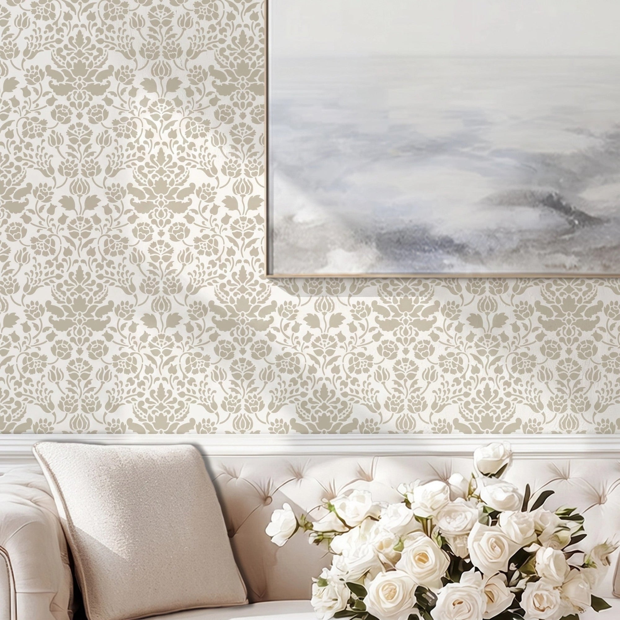 Floral damask wall stencil in a living room with a large landscape print hung on the wall, a cream-colored tufted sofa, and a vase of white roses, and natural light highlighting the light gray and taupe painted stencil on the wall.