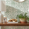 Floral damask wall stencil featuring a wooden floating shelf with two matching table lamps, a small potted plant, and decorative objects, and an round mirror, and natural light highlighting the green and white painted stencil on the wall.