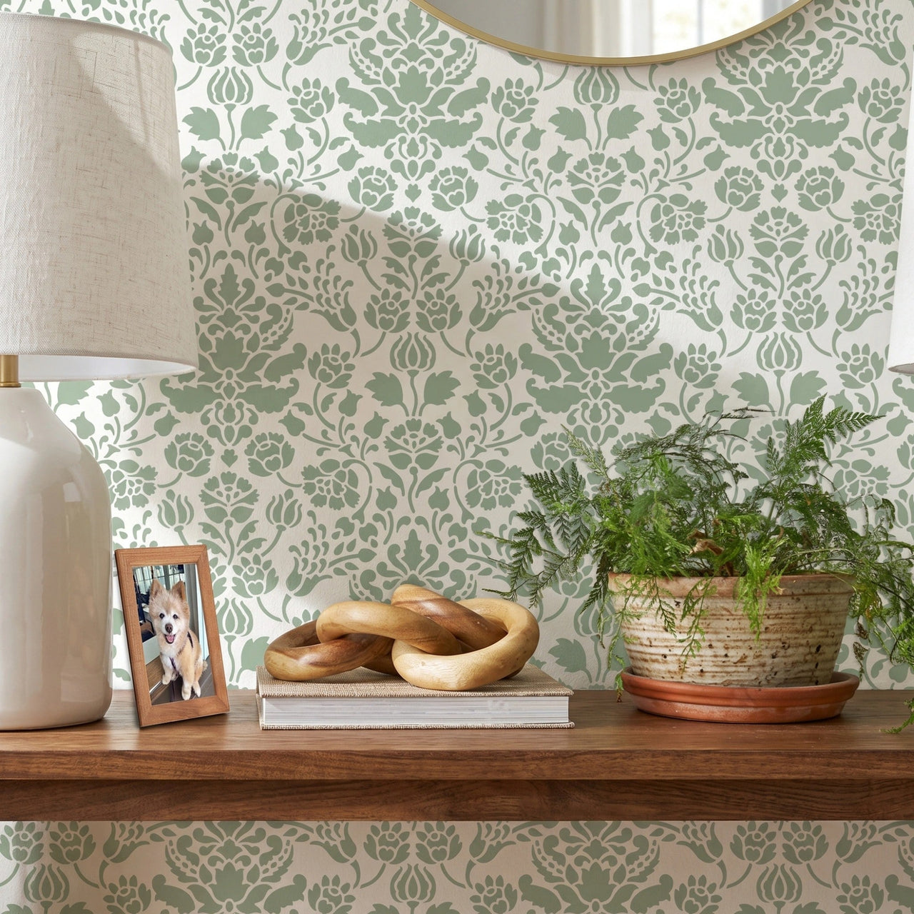 Floral damask wall stencil featuring a wooden floating shelf with two matching table lamps, a small potted plant, and decorative objects, and an round mirror, and natural light highlighting the green and white painted stencil on the wall.