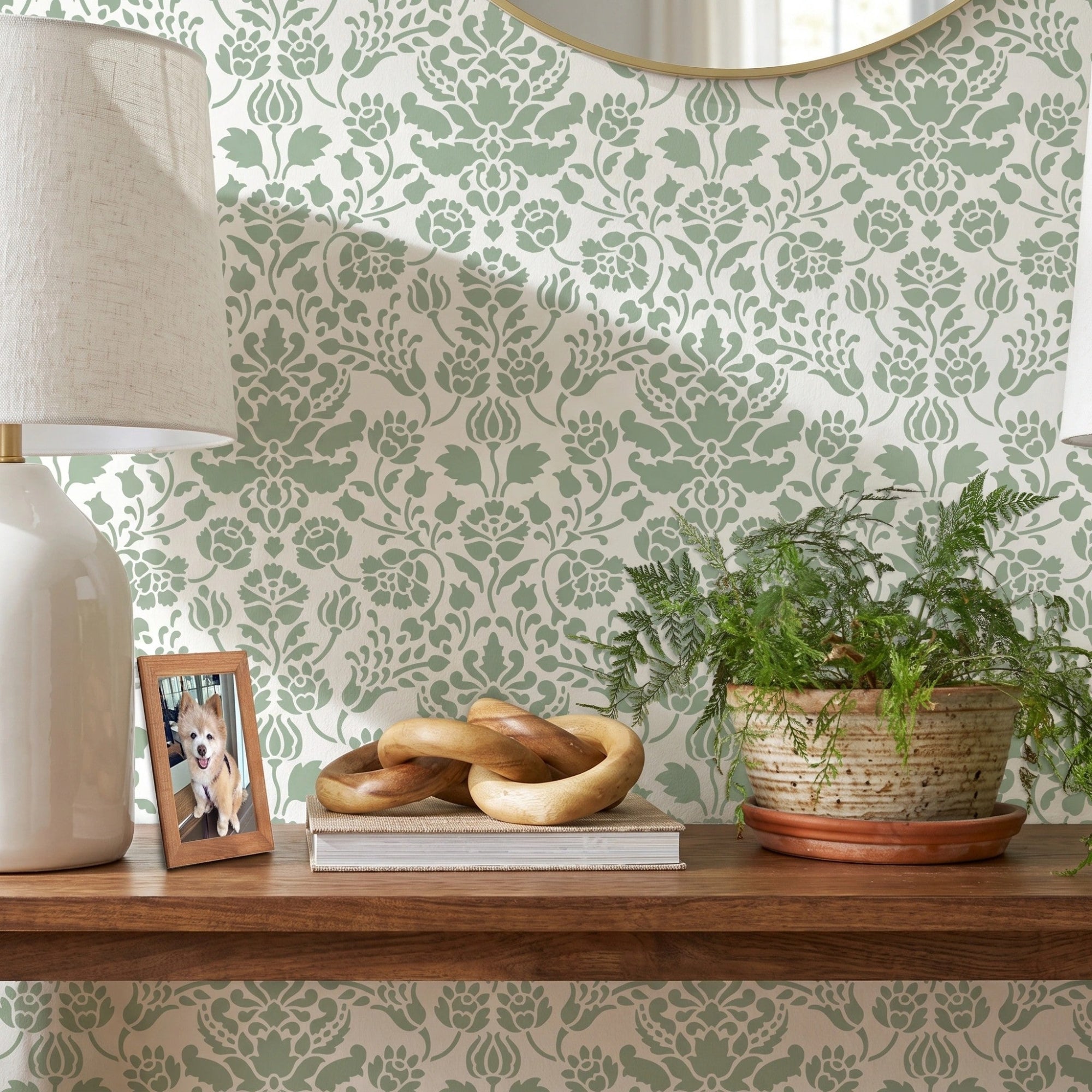 Floral damask wall stencil featuring a wooden floating shelf with two matching table lamps, a small potted plant, and decorative objects, and an round mirror, and natural light highlighting the green and white painted stencil on the wall.
