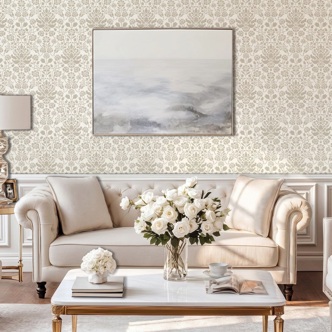 Floral damask wall stencil in a living room with a large landscape print, a cream-colored tufted sofa, a white marble coffee table, and a vase of white roses, and natural light highlighting the light gray and taupe painted stencil on the wall.