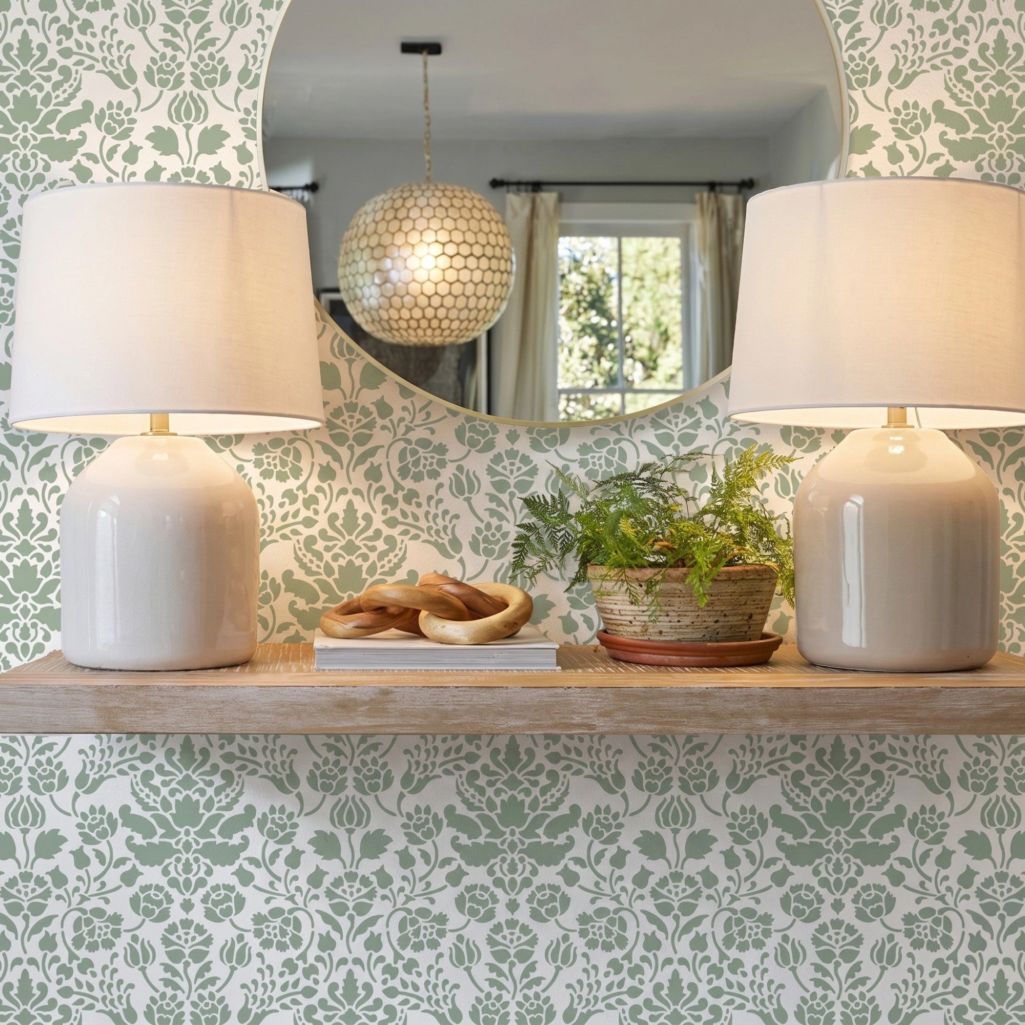 Floral damask wall stencil featuring a wooden floating shelf with two matching table lamps, a small potted plant, and decorative objects, and an round mirror, and natural light highlighting the green and white painted stencil on the wall.