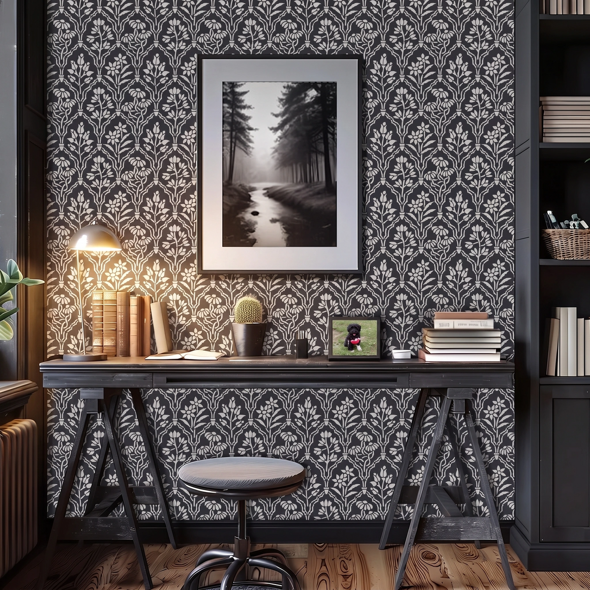 A styled home office scene featuring floral gallery wall stencil painted in light gray and charcoal grey paint, a framed black and white forest landscape print on the wall, a dark wood desk with a lamp, books, and a cactus, with a stool in the foreground.