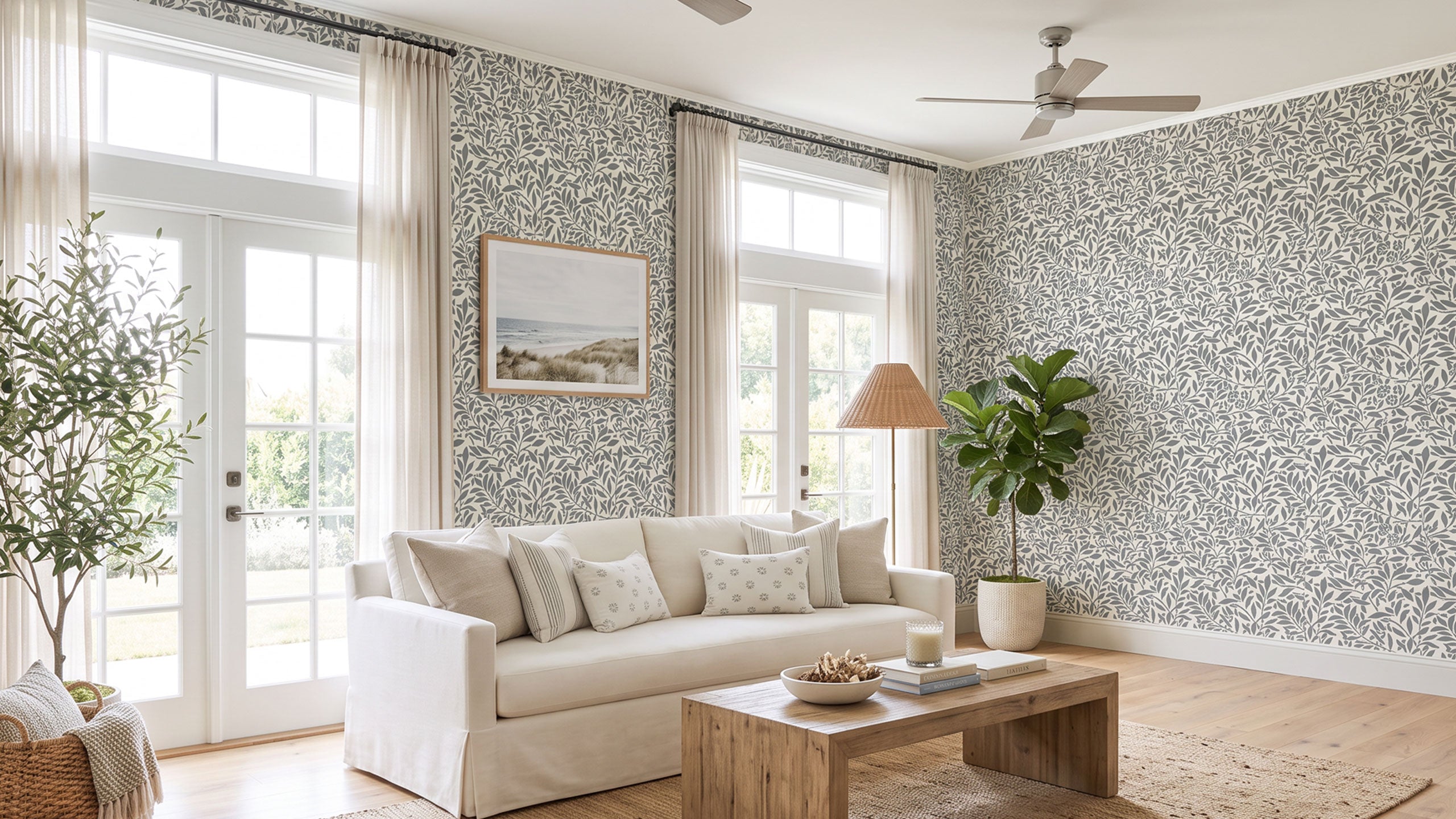 A bright, modern living room featuring a light beige slipcovered sofa, a wooden coffee table, and walls covered in a gray and white damask patterned wall stencil design. Large white French doors, sheer curtains, a floor lamp, and potted plants complete the scene.
