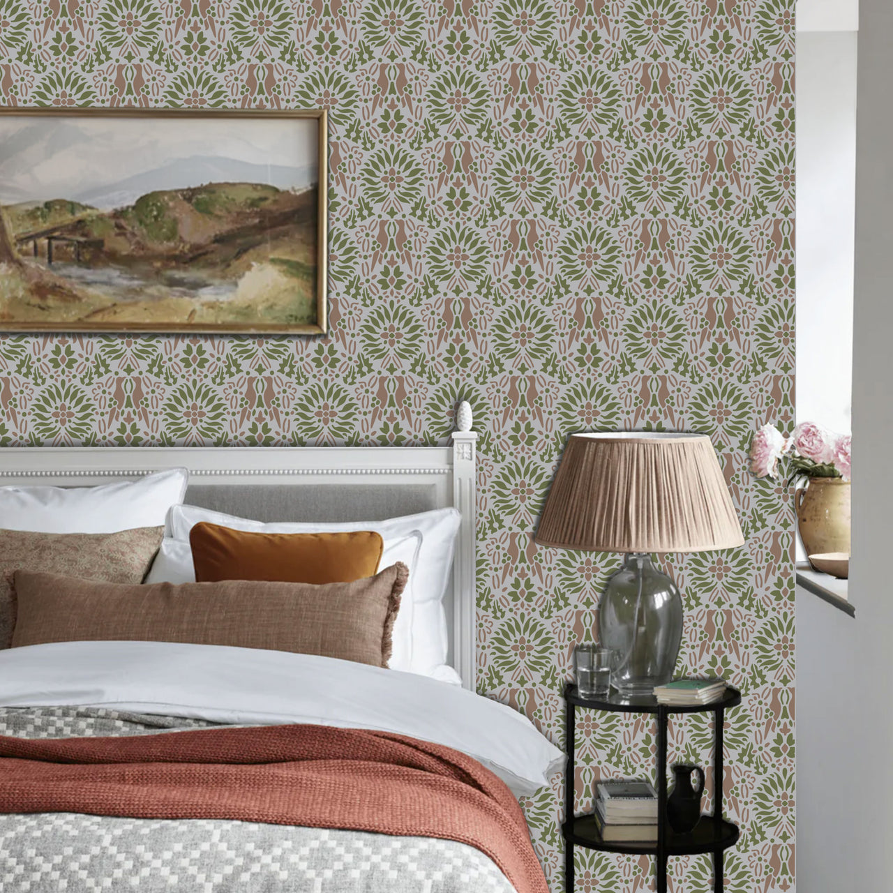 Morning dove stencil applied in a  bedroom corner, a white wooden bed frame, with a framed landscape painting on the wall and a pleated beige lamp on a glass table, and light highlighting the green, mauve and grey painted stencil on the wall.