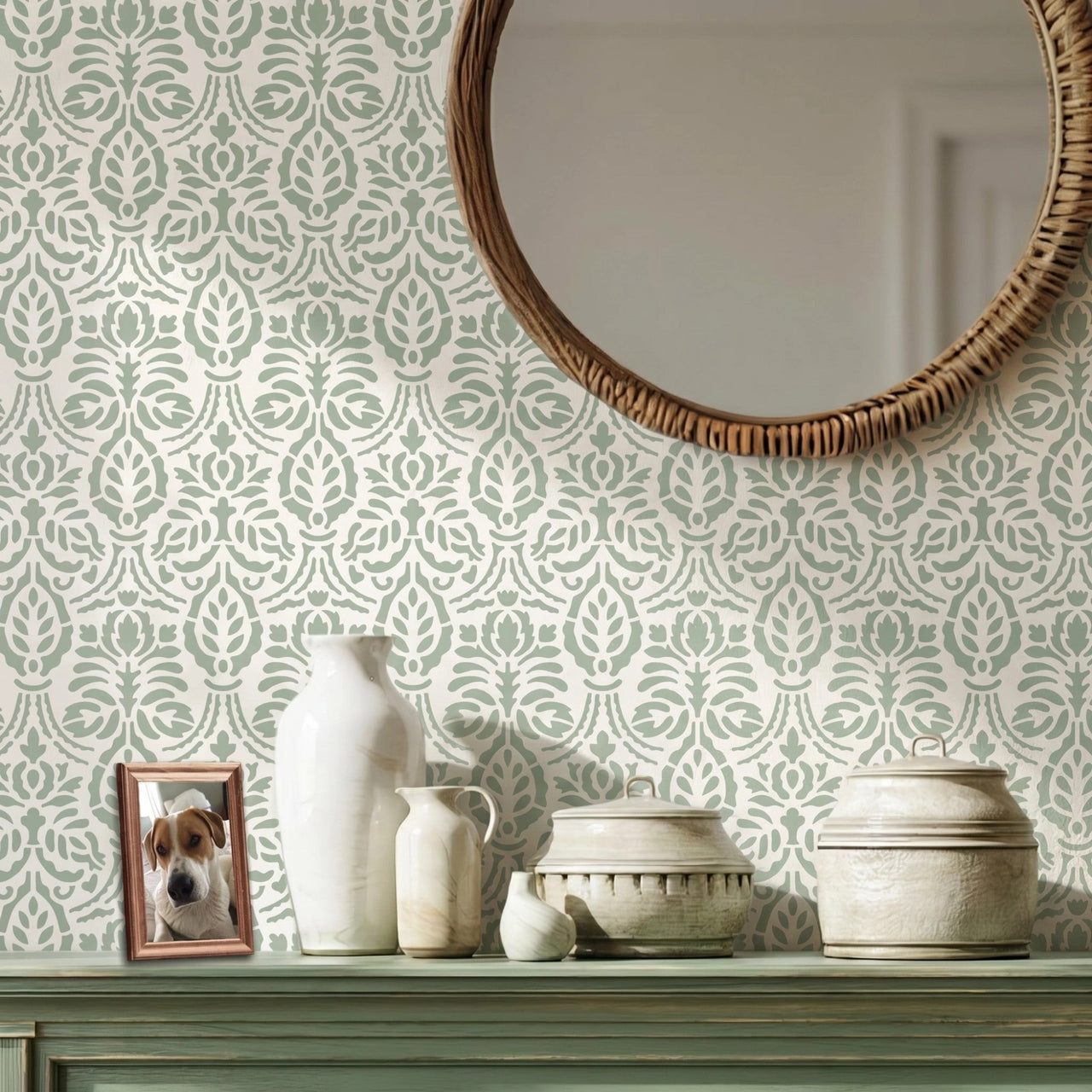 Spear Thistle wall stencil applied in a interior scene with a round woven mirror, a green console table, ceramic vases, and a framed picture of a dog, and natural light highlighting pale sage green and white painted floral stencil on the wall.