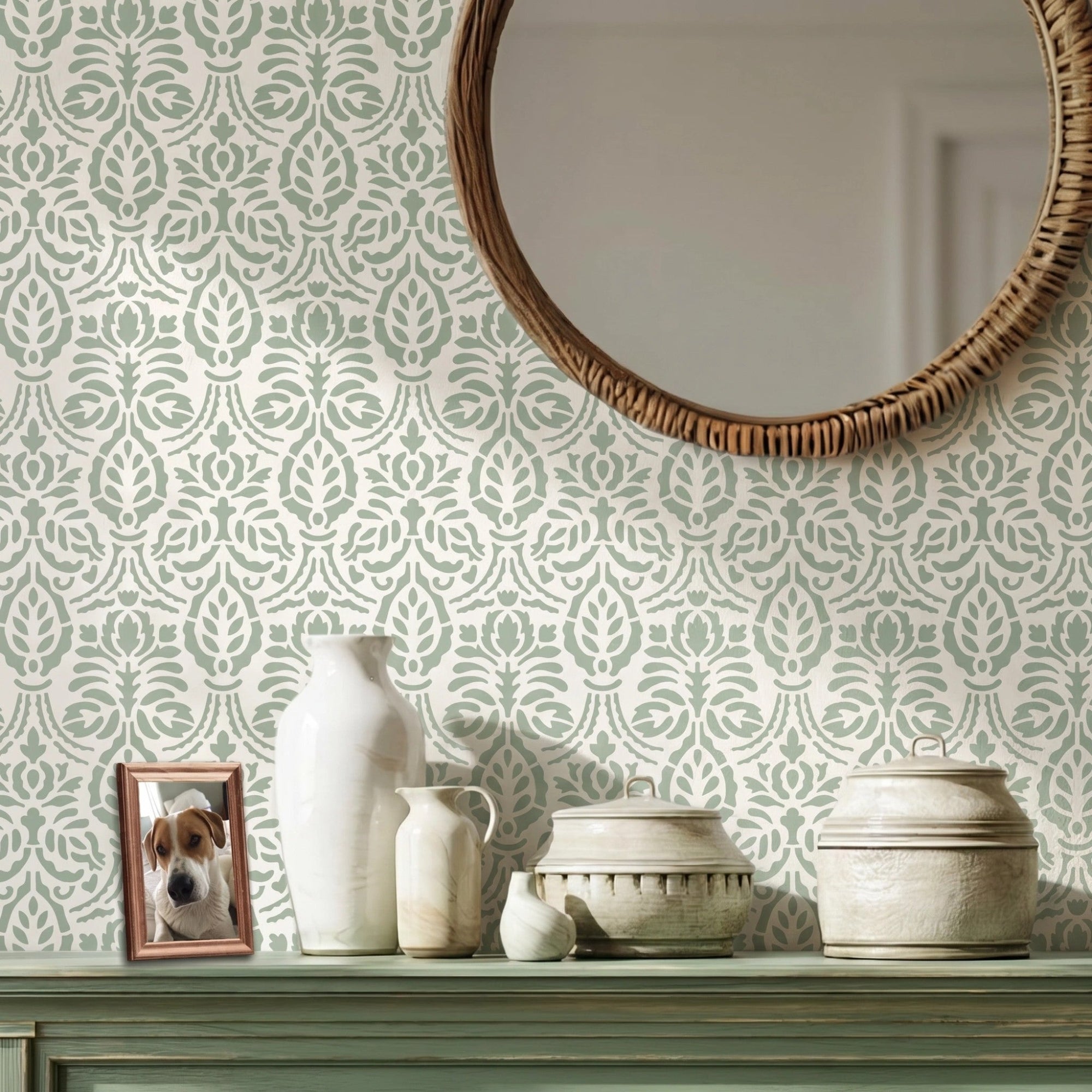 Spear Thistle wall stencil applied in a interior scene with a round woven mirror, a green console table, ceramic vases, and a framed picture of a dog, and natural light highlighting pale sage green and white painted floral stencil on the wall.