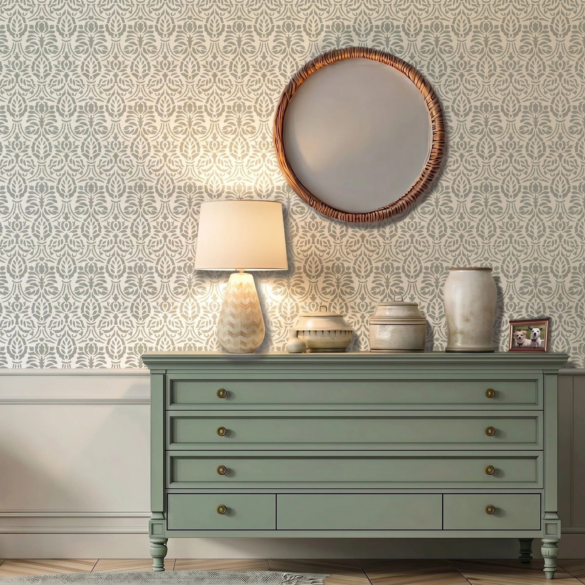 Spear Thistle wall stencil applied in a interior scene with a round woven mirror, a green console table, ceramic vases, and a framed picture of a dog, and natural light highlighting pale sage green and white painted floral stencil on the wall.