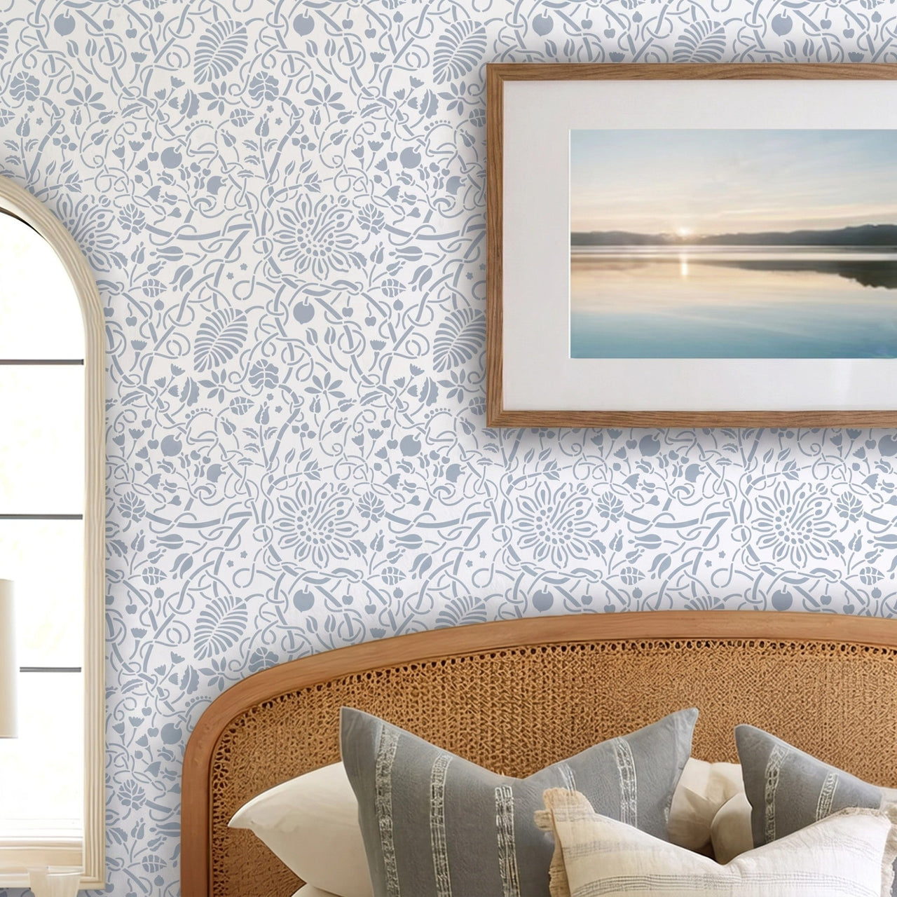 Swedish botanical wall stencil featured in a bedroom featuring, a framed landscape print on the wall, and a woven rattan headboard with neutral-toned pillows, and natural light highlighting the blue and white painted stencil on the wall.