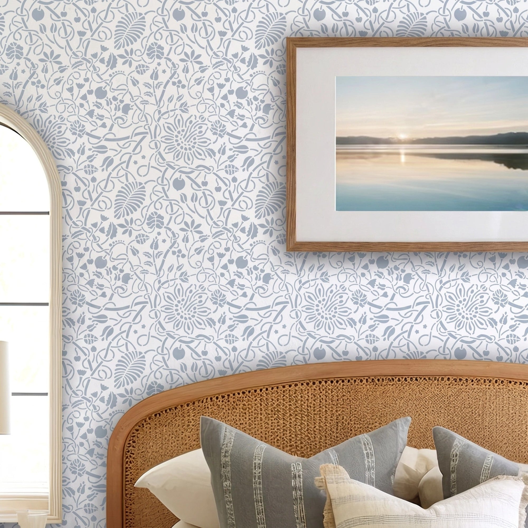 Swedish botanical wall stencil featured in a bedroom featuring, a framed landscape print on the wall, and a woven rattan headboard with neutral-toned pillows, and natural light highlighting the blue and white painted stencil on the wall.