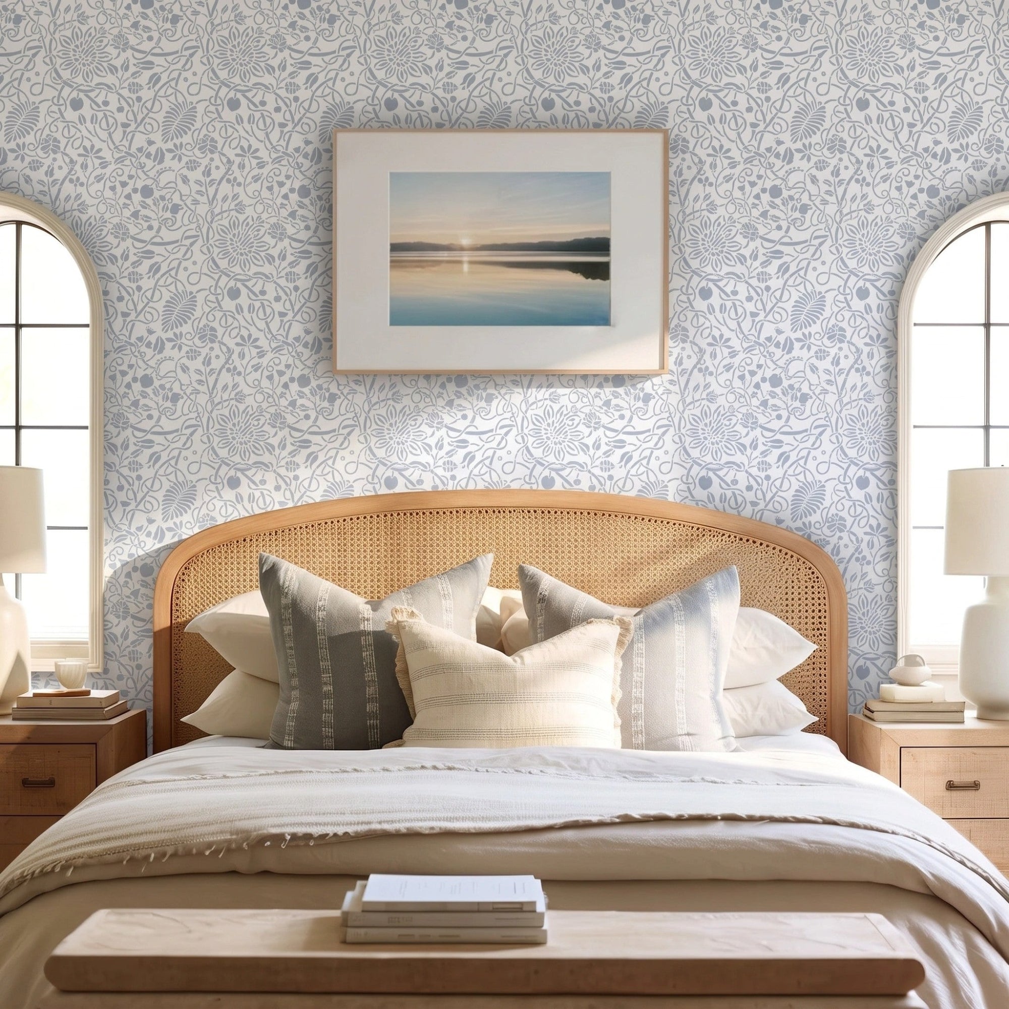 Swedish botanical wall stencil featured in a bedroom featuring, a framed landscape print on the wall, table lamps, a woven rattan headboard with neutral-toned pillows, and natural light highlighting the blue and white painted stencil on the wall.
