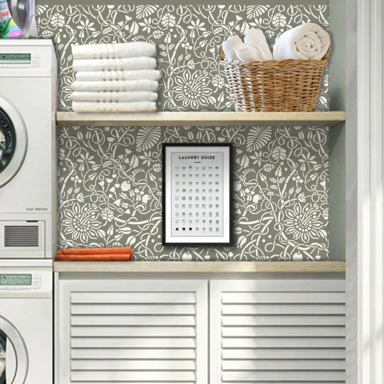 Swedish botanical wall stencil in a laundry machine in a entryway, wooden shelves, folded towels, a basket, and a framed image of laundry instruction symbols, with natural light highlighting the olive green and white painted stencil on the wall.