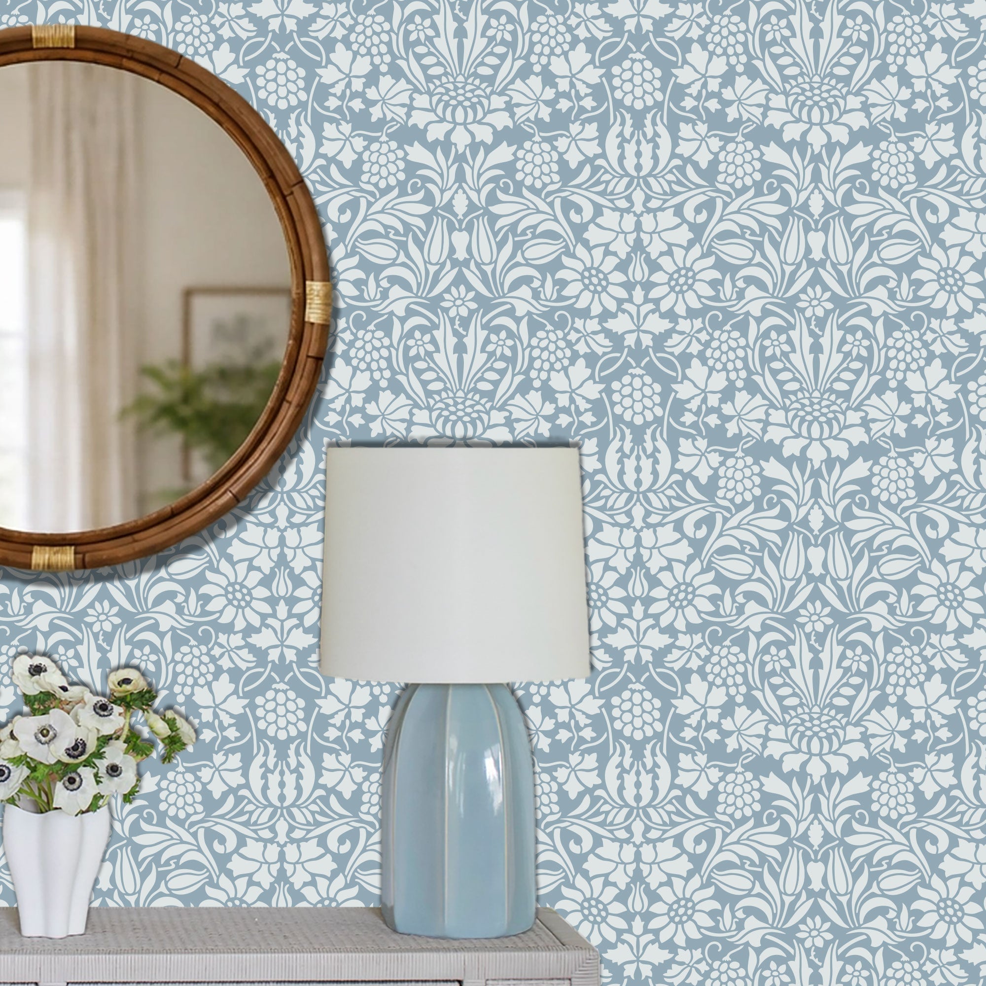 A styled interior scene showing a light blue and white damask wall stencil patterned wall behind a table lamp with a white drum shade and pale blue faceted base, plus part of a round wooden mirror and a white vase with flowers.