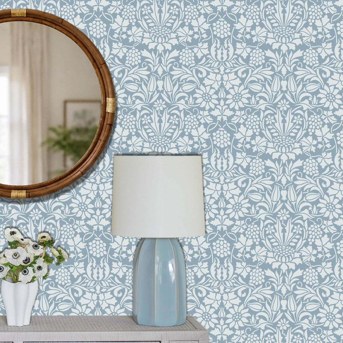 A styled interior scene showing a light blue and white damask wall stencil patterned wall behind a table lamp with a white drum shade and pale blue faceted base, plus part of a round wooden mirror and a white vase with flowers.