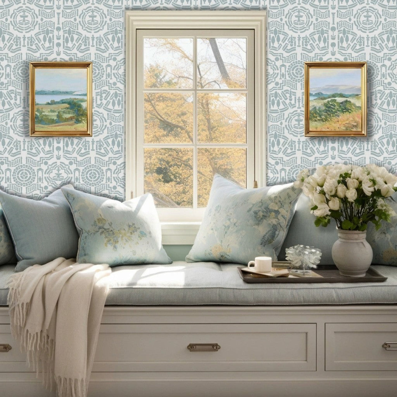 Topiary Garden wall stencil featuring a window seat cushion, two framed landscape paintings on the wall, and a vase of white flowers on a tray and a window with natural light highlighting the light blue and dark blue painted stencil on the wall.