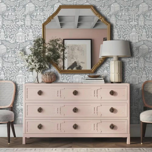 Video of Asian garden wall stencil applied in a styled interior corner featuring a pink dresser, a white table lamp with a ribbed cream base, a gold-framed mirror, two chairs, and natural light highlighting the gray and white painted stencil on the wall. Man enters the room with his dog, places his keys on the dresser and exits the room.