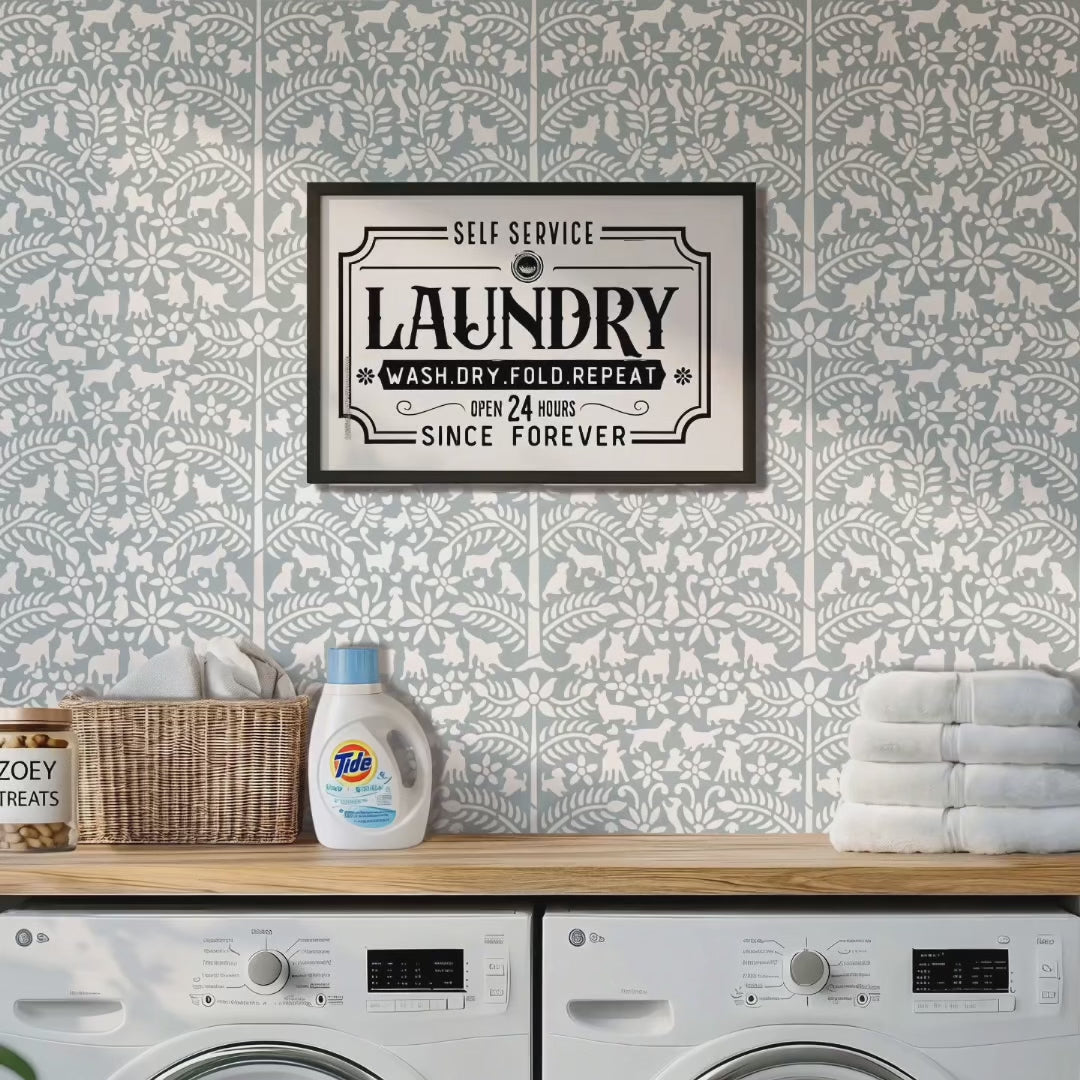 Dogs in the garden wall stencil applied in a laundry room with a washer and dryer, towels, detergent, basket and dog treats in a glass jar, and natural light highlights the light blue and white painted stencil on the wall. Woman enters the laundry room, gives a treat to the dog, picks up the folded towels and exits the room.