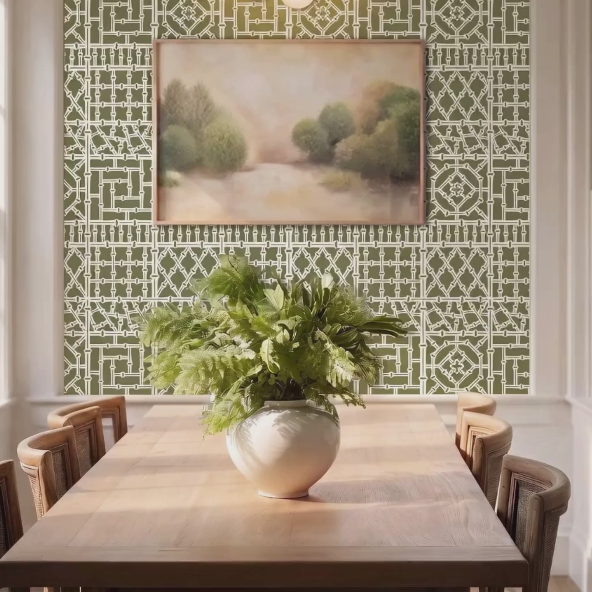Video of, bamboo wall stencil in a dining room, styled with a framed landscape painting and a green plant, and natural light highlighting the green and white painted stencil on the wall. woman enters and puts plates on the table.