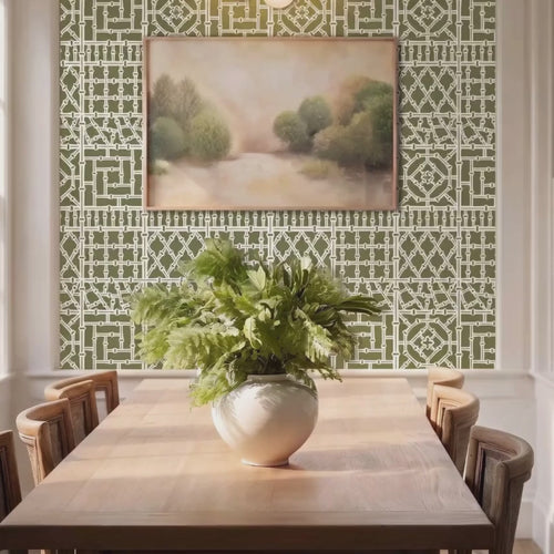 Video of, bamboo wall stencil in a dining room, styled with a framed landscape painting and a green plant, and natural light highlighting the green and white painted stencil on the wall. woman enters and puts plates on the table.