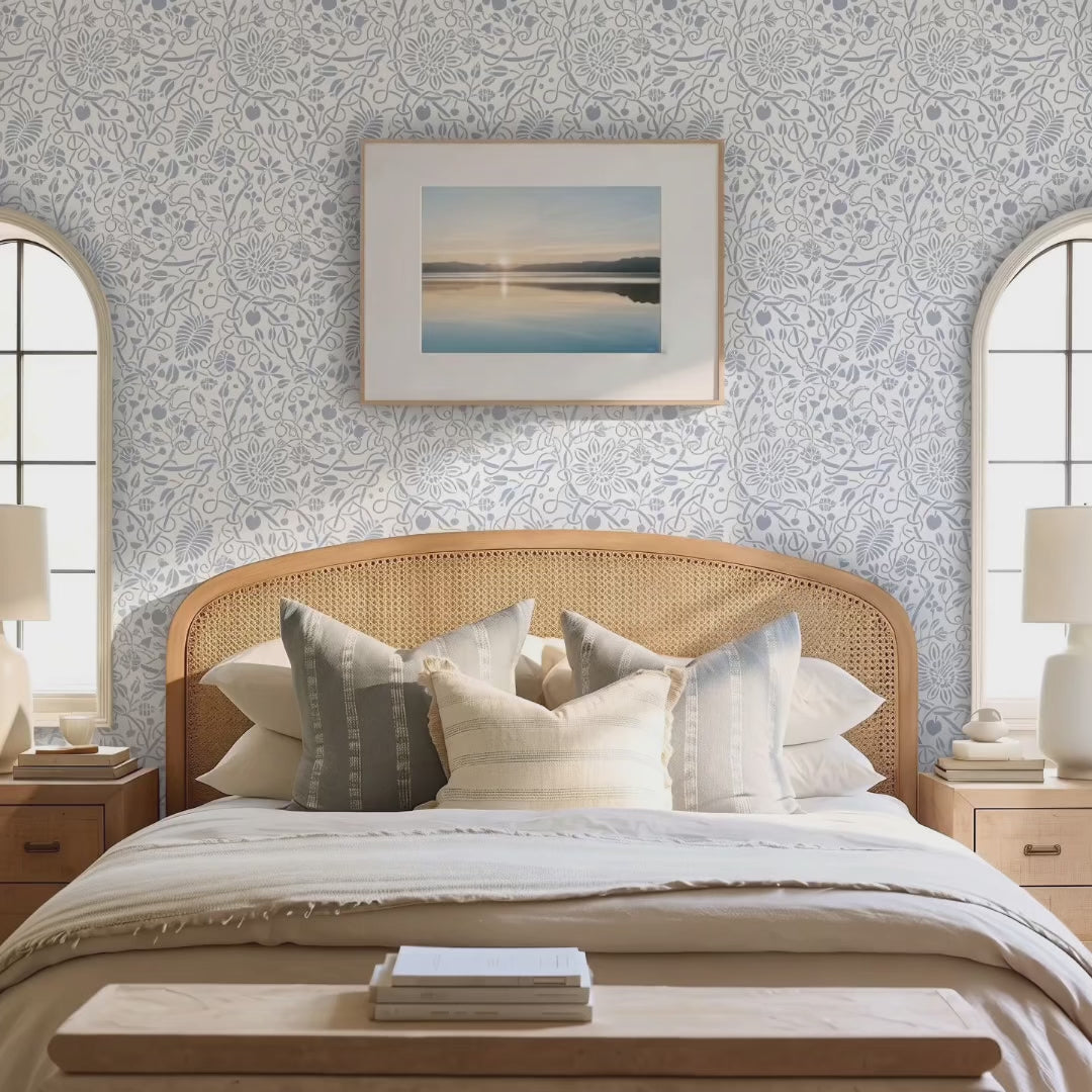 Video of Swedish botanical wall stencil featured in a bedroom featuring, a framed landscape print on the wall, table lamps, a woven rattan headboard with neutral-toned pillows, and natural light highlighting the blue and white painted stencil on the wall. Woman gets into bed and Pomeranian dog jumps onto the bed.