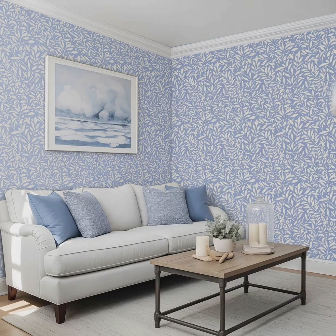 Video of hummingbird wall stencil on a wall above a sofa with blue and textured pillows, with a framed landscape print hanging on the wall, natural soft light highlighting the blue and white painted stencil on the wall. Two women walk into the living room, sit on the couch followed by a dog.