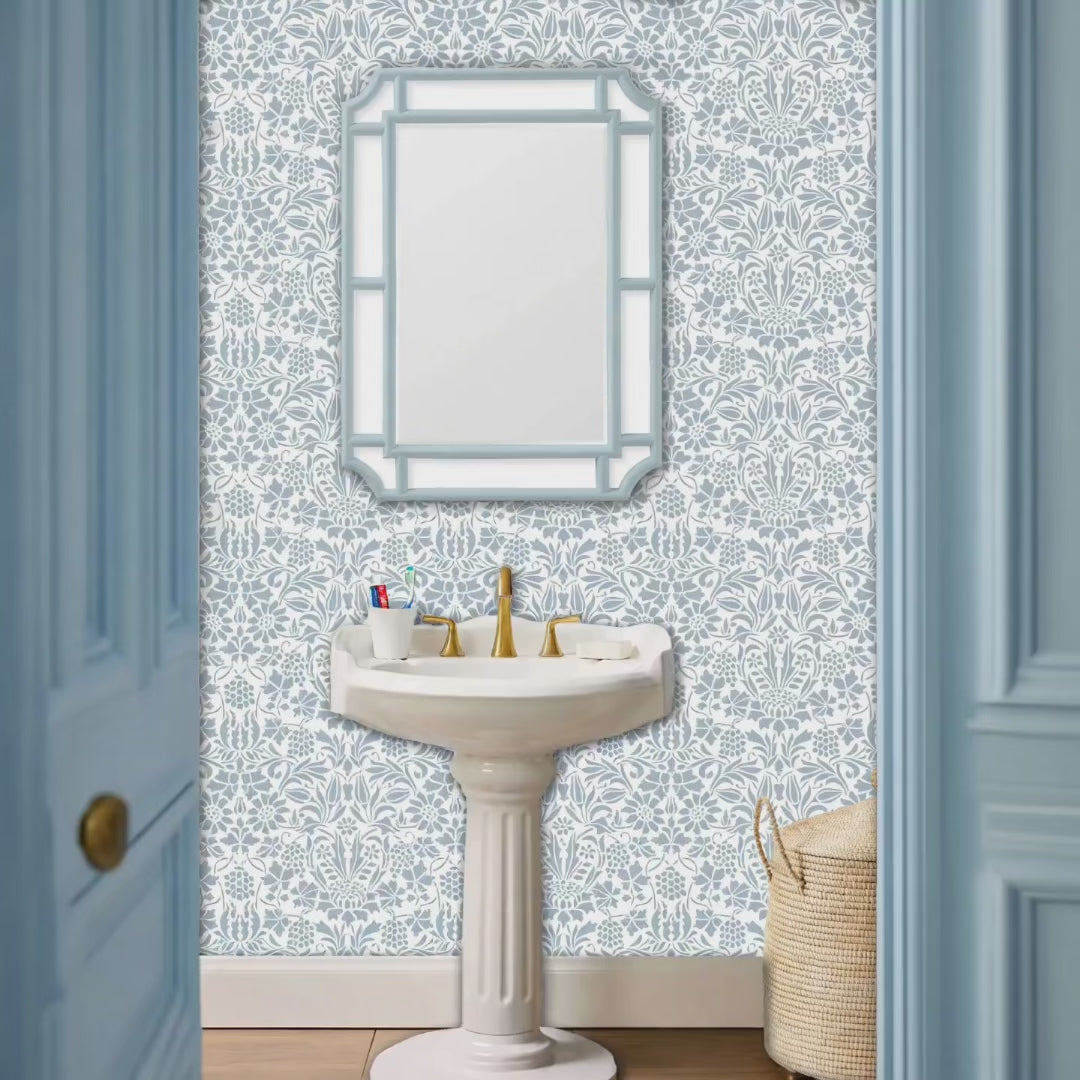 Video of thistle and vine wall stencil pattern applied in a powder room setting with a white sink, gold faucet, and a light blue-framed mirror, and natural light highlighting the blue and white painted stencil on the wall. Woman walks into powder room and closes the door.