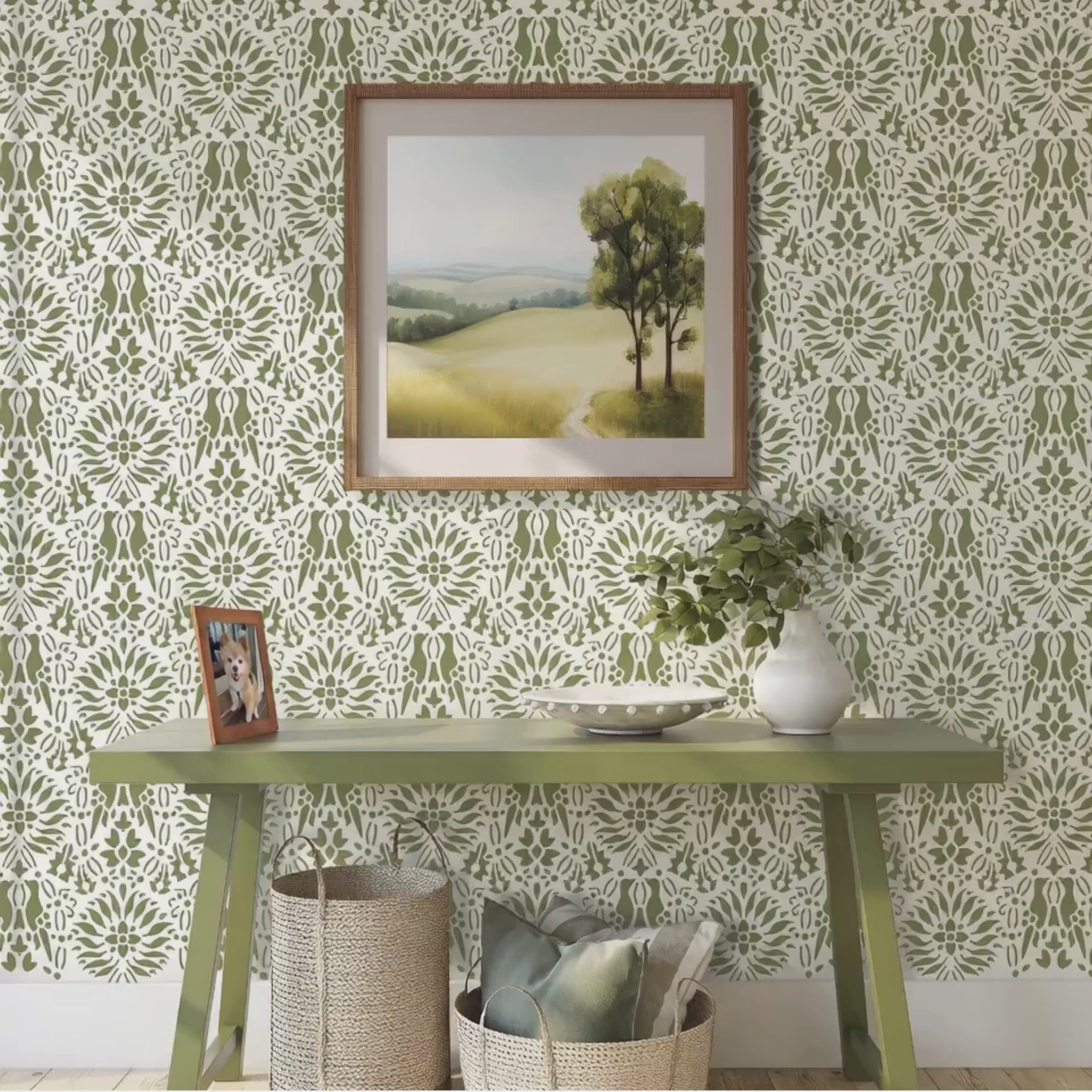 Video of Morning dove wall stencil applied in a styled room setting with a green console table, a framed landscape print, a white vase with greenery, and woven baskets, and soft natural light highlighting the green and white painted stencil on the wall. Woman walks in with her dog and places her keys on the table.