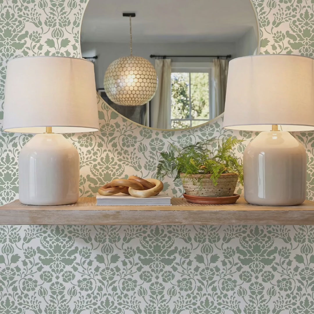 Video of floral damask wall stencil featuring a wooden floating shelf with two matching table lamps, a small potted plant, and decorative objects, and an round mirror, and natural light highlighting the green and white painted stencil on the wall. Woman enters carrying a watering can, waters the plant and exits the room.