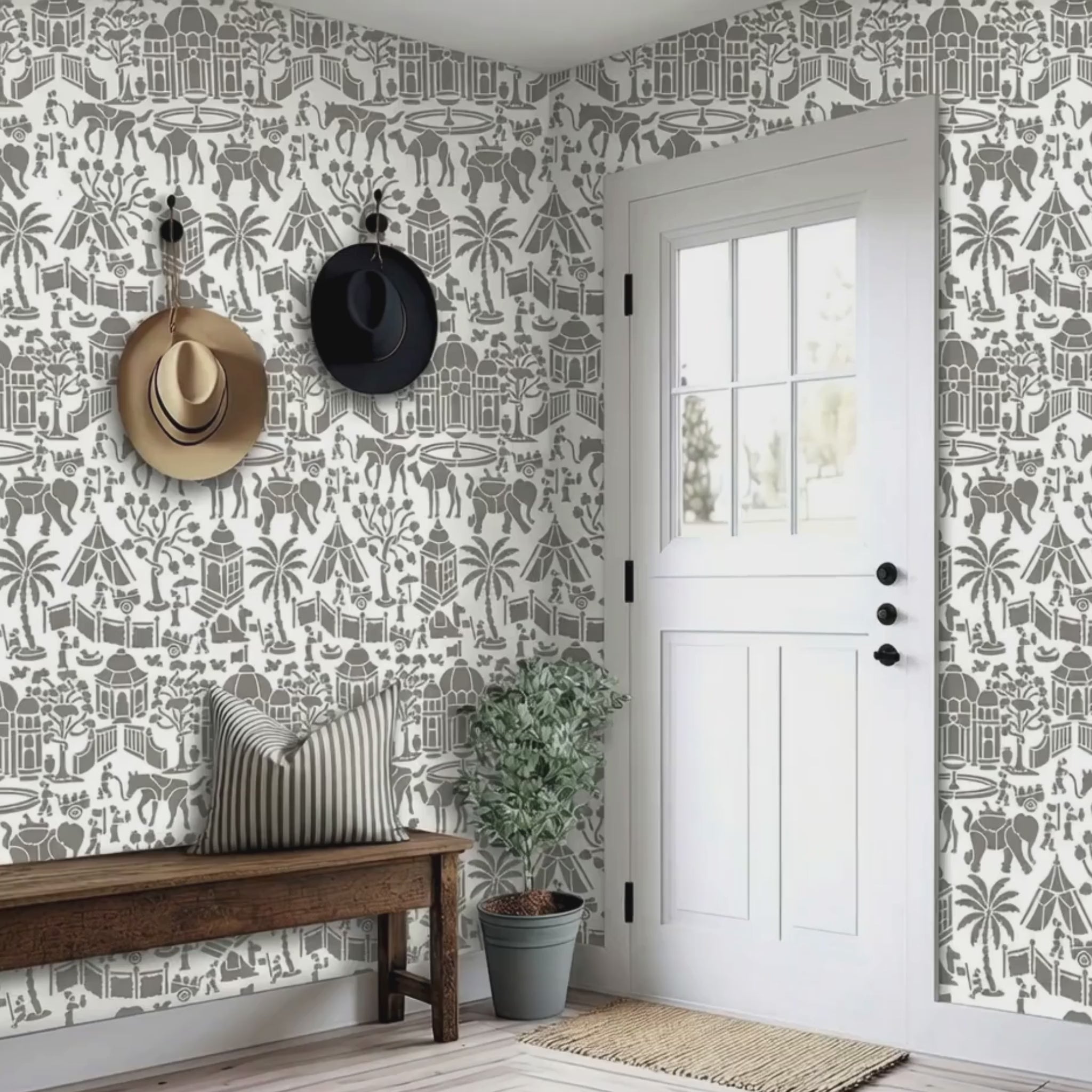 Video of indian village wall stencil applied in a entryway featuring a wooden bench, two hats hanging on the wall, a plant, a front door, and natural light highlighting the gray and white wall stencil painted on the wall. A woman and her door enter the room.