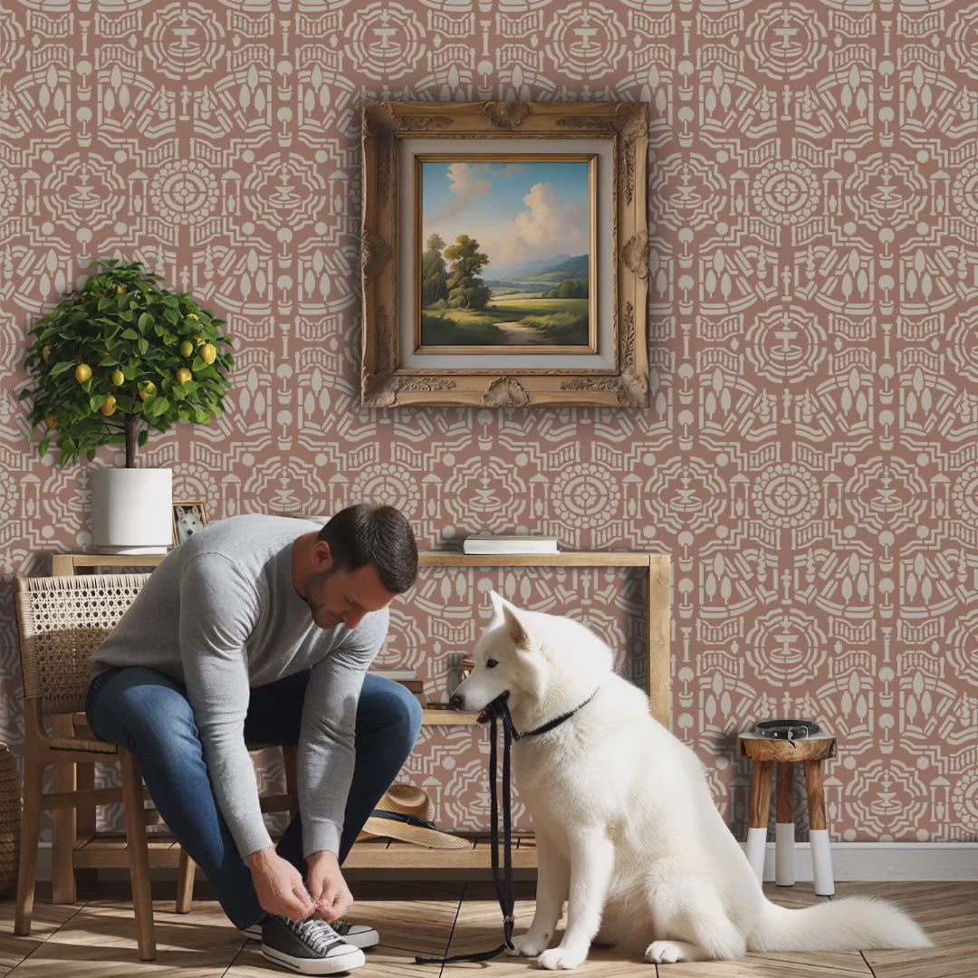 Video of topiary garden wall stencil applied in a hallway with a framed landscape painting hanging above a wooden table holding books and decorative objects, a chair, a plant and soft light highlighting the dusty rose and beige painted stencil on the wall. Man ties his shoes, retrieves the leash from the dog's mouth and leaves the room.